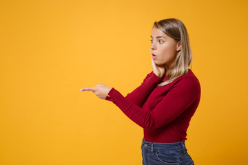 Side view of amazed young blonde woman girl in casual clothes posing isolated on yellow orange background. People lifestyle concept. Mock up copy space. Pointing index finger aside, put hand on cheek.