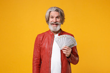 Cheerful elderly gray-haired mustache bearded man in red leather jacket isolated on yellow orange background. People lifestyle concept. Mock up copy space. Hold fan of cash money in dollar banknotes.
