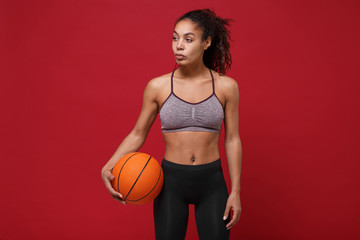 Attractive young african american sports fitness basketball player woman in sportswear working out isolated on red background. Sport exercises healthy lifestyle concept. Holding ball, looking aside.