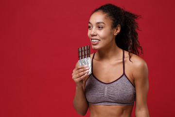 Smiling young african american sports fitness woman in sportswear working out isolated on red wall background in studio. Sport exercises healthy lifestyle concept. Hold chocolate bar looking aside.