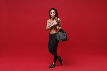 Smiling young african american fitness woman in sportswear posing working out isolated on red background studio portrait. Sport exercises healthy lifestyle concept. Holding sports bag with equipment.