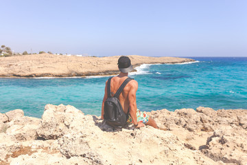 Fototapeta na wymiar Athletic young man in cap sitting around Natural Rock Bridge at Cape Greco near Ayia Napa in the evening light. Cyprus. Healthy lifestyle