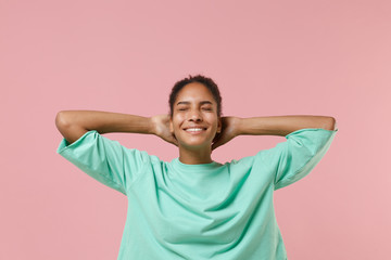 Smiling young african american woman girl in green sweatshirt posing isolated on pastel pink wall background. People lifestyle concept. Mock up copy space. Keeping eyes closed with hands behind head.