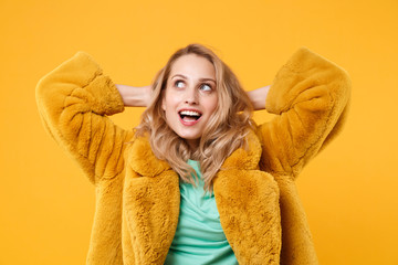 Pensive young blonde woman girl in yellow fur coat posing isolated on orange background, studio portrait. People emotions lifestyle concept. Mock up copy space. Holding hands behind head looking up.