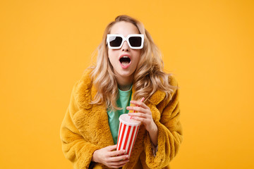 Shocked young blonde woman girl in yellow fur coat, dark sunglasses posing isolated on orange background in studio. People lifestyle concept. Mock up copy space. Holding plastic cup of cola or soda.