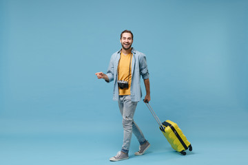 Laughing traveler tourist man in yellow summer casual clothes with photo camera isolated on blue background. Male passenger traveling abroad on weekends. Air flight journey concept. Holding suitcase.