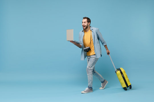 Excited traveler tourist man in yellow clothes with photo camera suitcase isolated on blue background. Male passenger traveling abroad on weekend. Air flight journey. Working on laptop booking hotel.