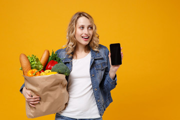 Pretty girl in denim clothes isolated on orange background. Delivery service from shop or restaurant concept. Hold brown craft paper bag for takeaway with food product mobile phone with empty screen.