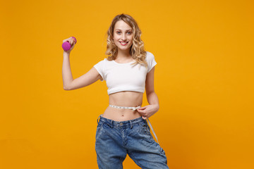 Smiling woman in white t-shirt isolated on orange background. Proper nutrition losing weight healthy sport lifestyle dieting. Wearing old big jeans measuring waist with measure tape hold dumbbell.