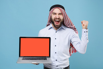 Joyful arabian muslim man in keffiyeh kafiya ring igal agal casual clothes isolated on pastel blue background. People religious concept. Hold laptop pc computer with blank screen doing winner gesture.