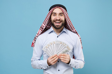 Cheerful bearded arabian muslim man in keffiyeh kafiya ring igal agal casual clothes isolated on pastel blue background. People religious lifestyle concept. Hold fan of cash money in dollar banknotes.