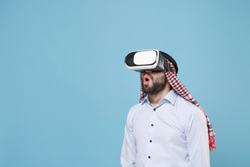 Shocked young bearded arabian muslim man in keffiyeh kafiya ring igal agal casual clothes isolated on pastel blue background. People religious lifestyle concept. Mock up copy space. Watch in headset.