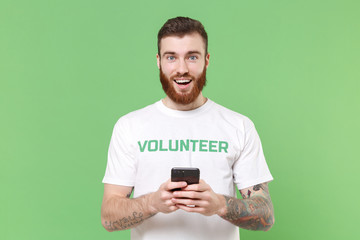 Excited young bearded man in white volunteer t-shirt isolated on pastel green background. Voluntary...