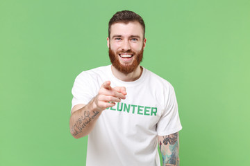 Laughing young bearded man in white volunteer t-shirt isolated on pastel green background in studio. Voluntary free work assistance help charity grace teamwork concept. Point index finger on camera.