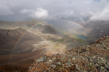 A full rainbow in a valley photographed from above from a mountain pass. Gloomy clouds in the sky. Below you can see the lake. Horizontal.