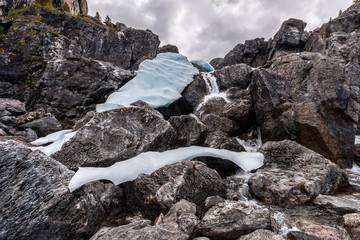 Huge boulders through which a mountain stream flows and the remains of ice on the stones. The sky is overcast. Horizontal.