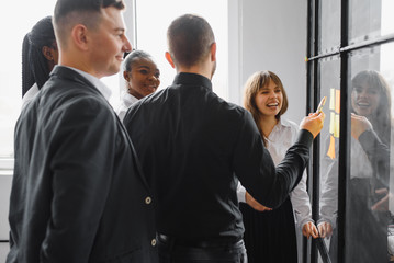 Happy mixed race colleagues watching smiling team leader marking task as done on colorful sticky notes of kanban agile glass window board at office. Diverse teammates satisfied with working process.