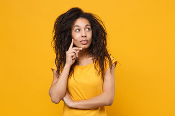 Pensive young african american woman girl in casual t-shirt posing isolated on yellow orange background in studio. People lifestyle concept. Mock up copy space. Put hand prop up on chin, looking up.