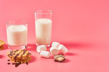 Two full glasses of fresh milk, yogurt or kefir near thin waffles, chocolate candies, marshmallow on pink background. Space for text