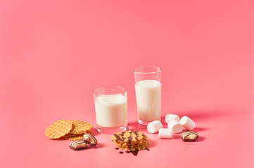 Two full glasses of fresh milk, yogurt or kefir near thin waffles, chocolate candies, marshmallow on pink background. Space for text