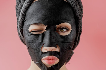 Young woman apply black cosmetic fabric facial mask on pink background. Face peeling mask with charcoal, spa beauty treatment, skincare, cosmetology. Close up