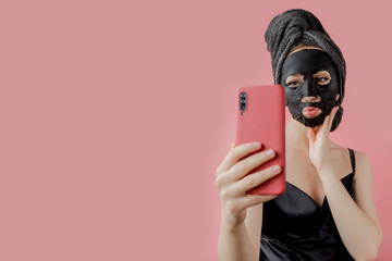 Young woman apply black cosmetic fabric facial mask and phone in hands on pink background. Face peeling mask with charcoal, spa beauty treatment, skincare, cosmetology. Close up