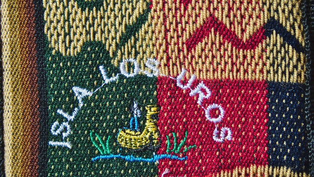 Embroidery texture with the letters "Isla los Uros" with a golden raft on a lake, the texture shows portions of green, red, gold, brown, yellow, orange and black fabric.