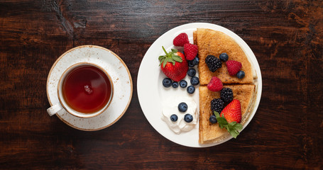Pancakes with berries and sour cream on a dark wooden background. Sweet homemade pancakes and a cup of tea on the table. Top view.