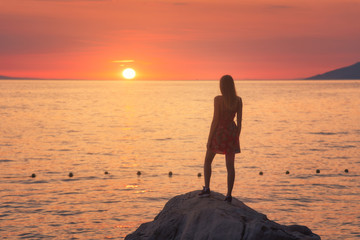 Young woman standing on the rock in the sea against mountains and colorful orange sky at sunset in summer. Landscape with alone slim girl on the stone, seashore and sunlight. Travel