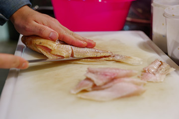 Close up view of Chef'hand use knife for cut and slice red snapper fish on white chopping block and sliced pieces of fish fillet, on countertop in kitchen.