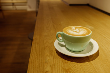 View latte art with heart pattern on top of cappuccino coffee on pale green ceramic cup on wooden counter inner coffee shop.