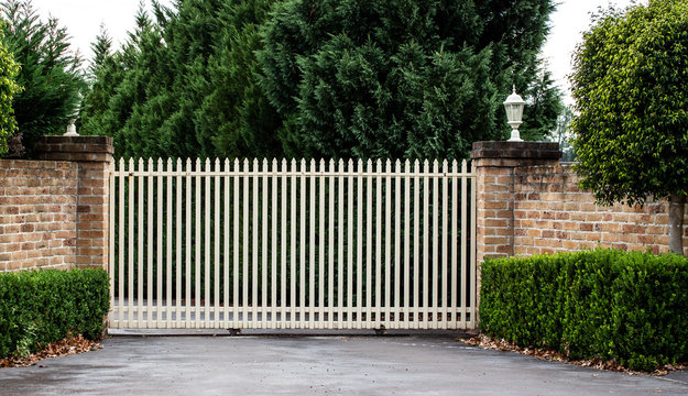 Metal wrought iron driveway property entrance gates set in brick fence, garden trees in background