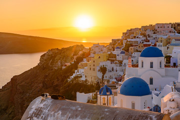 Blue domed churches and traditional white houses facing Aegean Sea with warm sunset light in Oia, Santorini, Greece