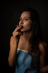 Fototapeta premium Portrait of Asian/Japanese/Korean/Indian brunette young girl in blue bath towel smoking cigarette in black studio copy space background. Indian lifestyle and fashion photography
