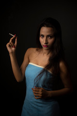 Fototapeta premium Portrait of Asian/Japanese/Korean/Indian brunette young girl in blue bath towel smoking cigarette in black studio copy space background. Indian lifestyle and fashion photography