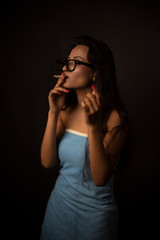 Portrait of Asian/Japanese/Korean/Indian brunette young girl in blue bath towel  and spectacles smoking cigarette in black studio copy space background. Indian lifestyle and fashion photography