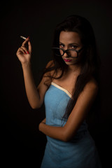 Portrait of Asian/Japanese/Korean/Indian brunette young girl in blue bath towel  and spectacles smoking cigarette in black studio copy space background. Indian lifestyle and fashion photography