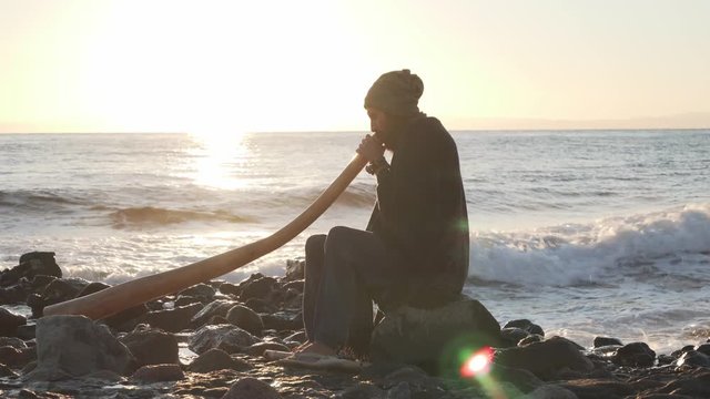 View of man sitting on seashore in sunlight and playing didjireedoo instrument