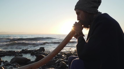 Bearded man playing his didgeridoo Australian instrument in sunshine at seashore