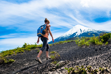 Young woman hiker walks on the loose rocky trail with snow capped volcano on the background. Trekking to volcano of Osorno in Chile