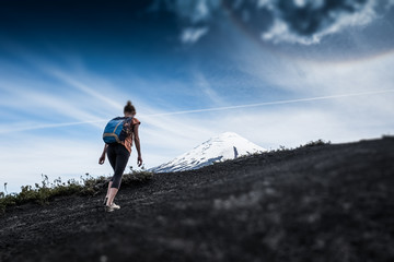 Obraz premium Young woman hiker walks on the deserted loose rocky trail on the mountain slope with snow capped volcano of Osorno on the background in Chile. Tilt shift effect applied