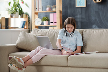 Teenage girl with headphones on neck using internet resources on laptop while doing homework in living room