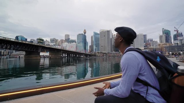 Black male tourist sits down on a bench and admires Darling Harbour, Pyrmont bridge and the city skyline on a quiet, overcast Sydney day. 