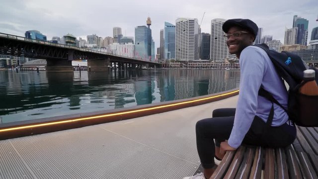 African american male sitting on a bench smiles at the camera with Darling Harbour and the Sydney skyline visible in the background. 