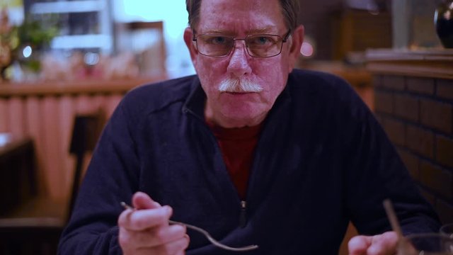 Handsome senior man sits in restaurant and chews food, looks at camera