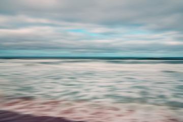 Abstract  seascape with blurred panning motion. Overcast day, ocean waves, and cloudy sky. Image displays blue and light pink split-toned color scheme.