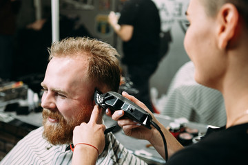  In a barber shop, a young woman cuts a guy with red hair and a red beard