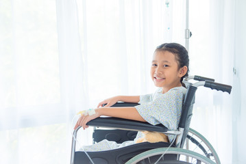 Asian sick girl with intravenous set on left hand sitting on wheel chair and smiles at hospital.