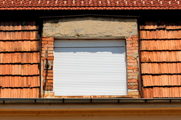 Fototapeta na wymiar Closed new plastic window blinds covering double window mounted on side of suburban family house below renovated roof surrounded with dilapidated cracked roof tiles