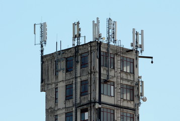 Concrete tall dilapidated old rarely used industrial building with multiple windows and metal steps on one side surrounded with densely mounted cell phone antennas and transmitters on top on clear blu